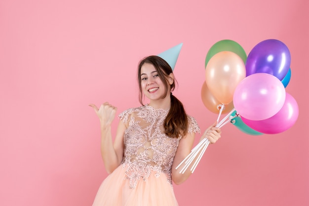 gelukkig feestmeisje met feestmuts met ballonnen die met de vinger iets op roze wijzen