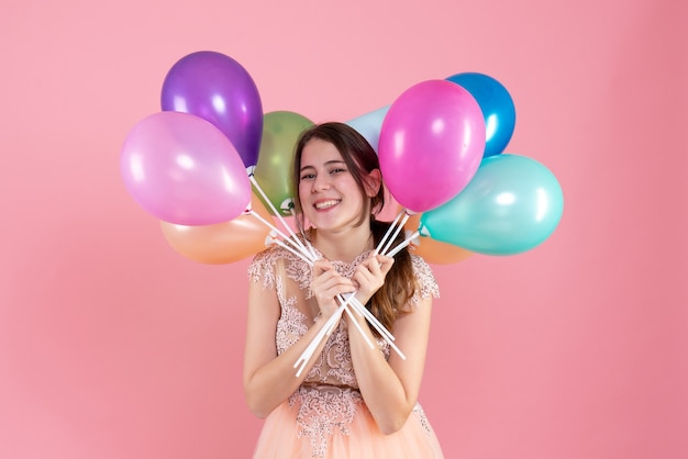 gelukkig feestmeisje met feestmuts met ballonnen in de buurt van haar gezicht op roze