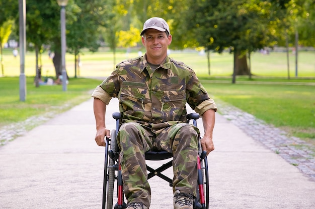 Gelukkig gehandicapte militaire man in rolstoel dragen camouflage uniform, verplaatsen op voetpad in stadspark. Vooraanzicht. Veteraan van oorlog of handicap concept