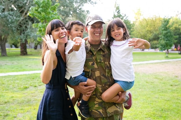 Gelukkig gezin paar en twee kinderen poseren in park. Militaire man met kinderen in de armen, zijn vrouw hen knuffelen en zwaaien. Gemiddeld schot. Familiereünie of het concept van thuiskomst