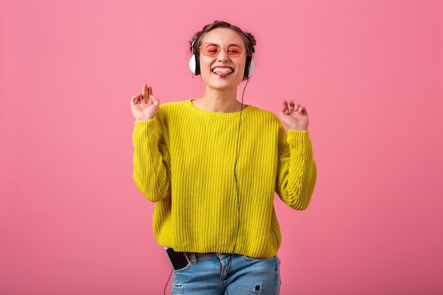 Gelukkig grappige vrouw luisteren naar muziek in koptelefoon gekleed in hipster kleurrijke stijl outfit geïsoleerd op roze muur, gele trui en zonnebril dragen, plezier tonen tong