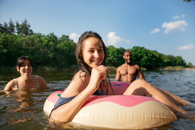 Gelukkig groep vrienden plezier tijdens het lachen en zwemmen in de rivier. Vrolijke mannen en vrouwen met rubberen ringen als een donut aan de rivier in zonnige dag. Zomer, vriendschap, toevlucht, weekendconcept.