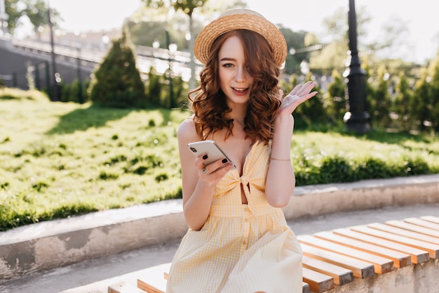 Gelukkig innemend meisje met golvend rood haar, zittend op een bankje met telefoon. outdoor portret van enthousiaste gember vrouw ochtend doorbrengen in park.