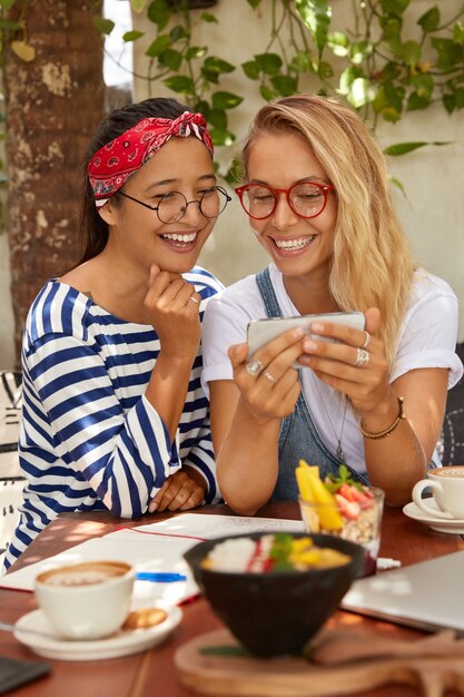 Gelukkig interraciale jonge meisjes lachen om grappige foto's, bekijken op slimme telefoon, veel plezier samen