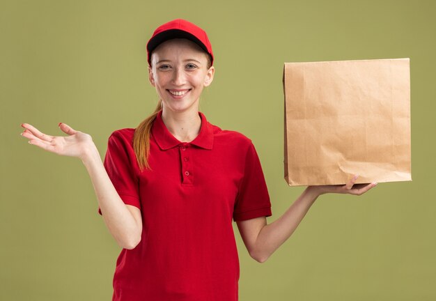 Gelukkig jong bezorgmeisje in rood uniform en pet met pakket glimlachend zelfverzekerd met arm uit arm