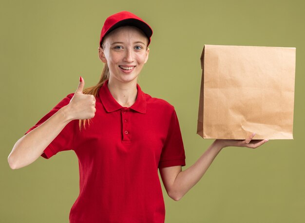 Gelukkig jong bezorgmeisje in rood uniform en pet met pakket glimlachend zelfverzekerd met duim omhoog