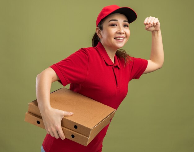 Gelukkig jong bezorgmeisje in rood uniform en pet rush running voor het leveren van pizzadozen voor klant over groene achtergrond