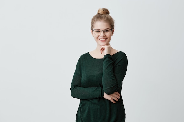 Gelukkig jong blonde wijfje dat haar haar heeft dat in knoop wordt gebonden die grote glazen draagt en het groene sweater stellen tegen de grijze hand van de muurholding onder haar kin die brede glimlach heeft