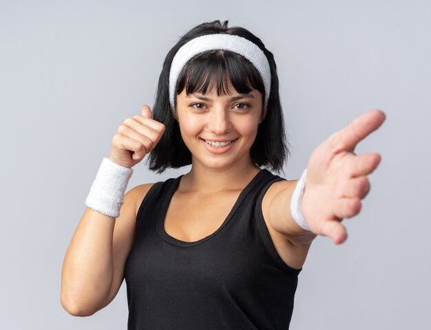 Gelukkig jong fitnessmeisje dat een hoofdband draagt en naar een camera kijkt die glimlacht en duimen laat zien en hier een gebaar met de hand maakt