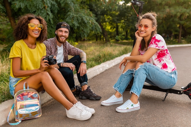 Gelukkig jong gezelschap van lachende vrienden zitten in park op gras met elektrische kick scooter, man en vrouw samen plezier