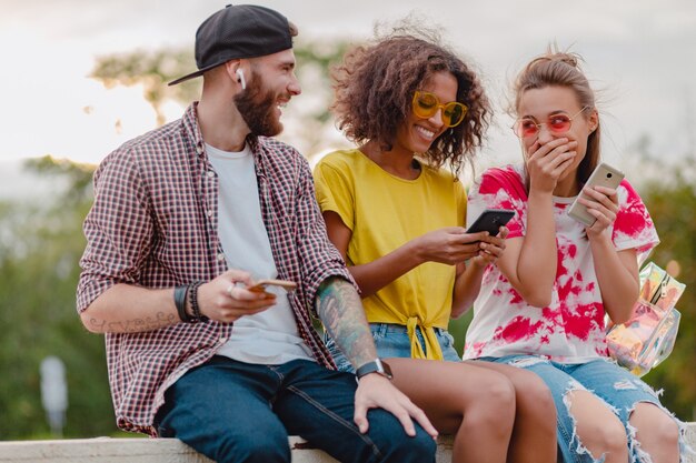 Gelukkig jong gezelschap van lachende vrienden zitten park met behulp van smartphones, man en vrouw met plezier