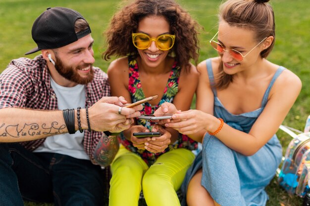 Gelukkig jong gezelschap van lachende vrienden zitten park met behulp van smartphones, man en vrouw samen plezier