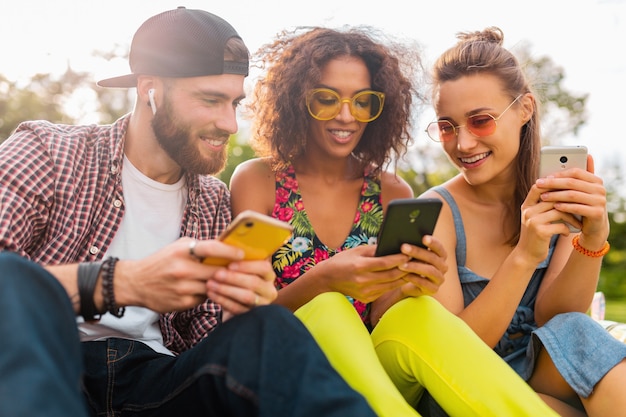 Gelukkig jong gezelschap van lachende vrienden zitten park met behulp van smartphones, man en vrouw samen plezier