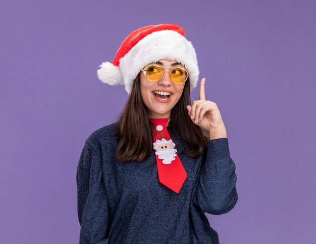 Gelukkig jong Kaukasisch meisje in zonnebril met kerstmuts en santa stropdas wijst omhoog kijkend naar kant geïsoleerd op paarse muur met kopie ruimte