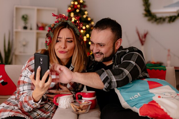 gelukkig jong koppel man en vrouw zittend op een bank met kopjes thee met plezier met smartphone in ingerichte kamer met kerstboom op de achtergrond