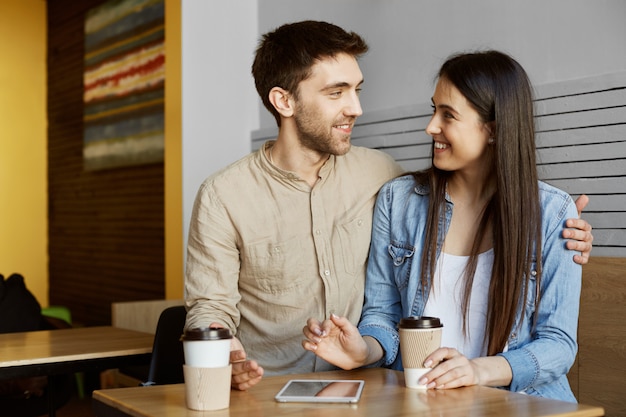 Gratis foto gelukkig jong koppel van twee stijlvolle studenten zitten in de cafetaria, koffie drinken, glimlachen, knuffelen en praten over hun leven.