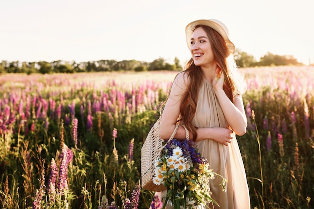 Gratis foto gelukkig jong meisje die op bloemgebied bij zonsondergang lopen. het dragen van strohoed en zak vol bloemen.