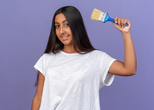 Gelukkig jong meisje in een wit t-shirt met een verfkwast die naar een camera kijkt die vrolijk over blauw staat te glimlachen