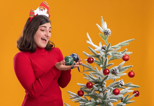 Gelukkig jong meisje in Kerstmissweater die grappige hoofdband dragen die x-mas ballen naast een kerstboom houden over oranje achtergrond