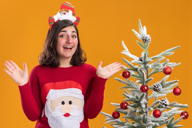 Gratis foto gelukkig jong meisje in kerstmissweater die grappige hoofdband met glimlach op gezicht dragen die zich naast een kerstboom over oranje muur bevindt