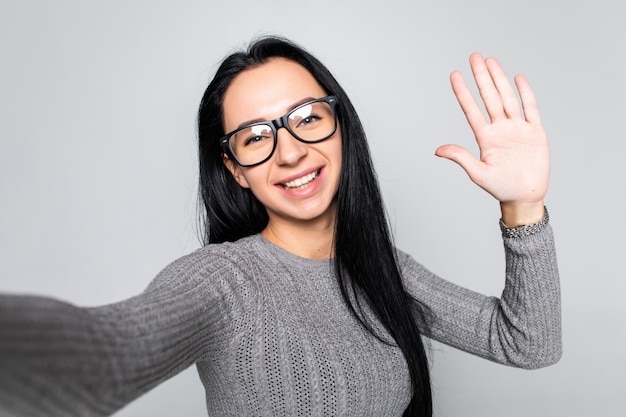 Gelukkig jong meisje zwaaiende hand naar camera terwijl het nemen van een selfie met mobiele telefoon geïsoleerd over grijze muur