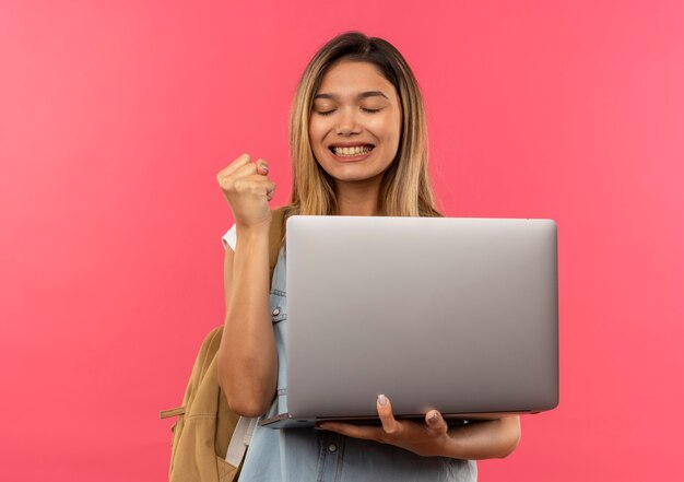 Gelukkig jong mooi studentenmeisje die laptop van de achterzakholding dragen en balde vuist met gesloten ogen die op roze met exemplaarruimte worden geïsoleerd