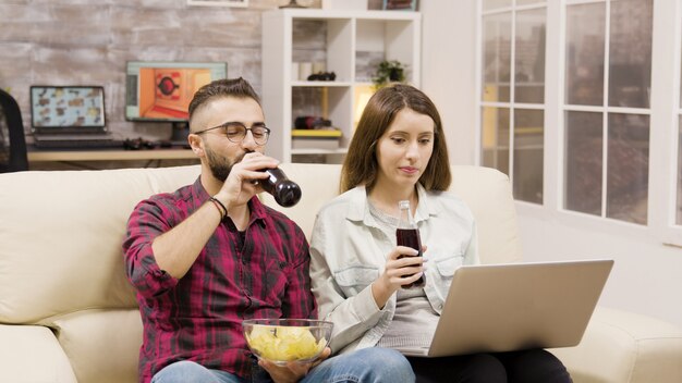 Gelukkig jong stel doet online winkelen op laptop. Paar zittend op de bank frisdrank drinken en chips eten.