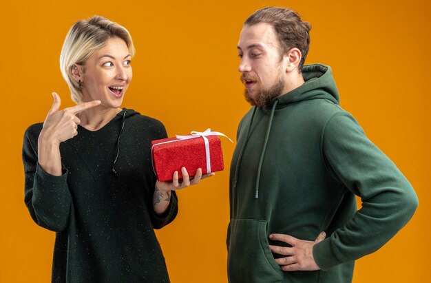 gelukkig jong stel in vrijetijdskleding man en glimlachende vrouw met heden wijzend met wijsvinger naar haar vriendje vieren Valentijnsdag staande over oranje muur