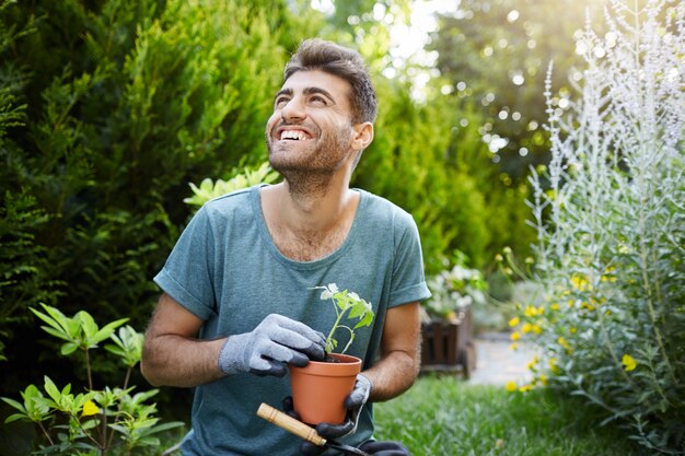 Gelukkig jonge aantrekkelijke bebaarde Kaukasische mannelijke tuinman in blauw t-shirt en handschoenen glimlachen, bloempot met groene spruit in handen houden, opzij kijken met opgewonden gezichtsuitdrukking