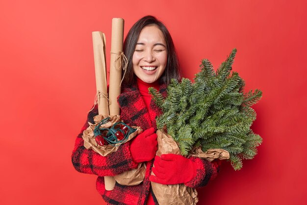 Gelukkig jonge Aziatische vrouw lacht vreugdevol houdt groene vuren takken verpakt in papier retro slinger keert terug van kerstmarkt bereidt zich voor op vakantie draagt winterkleren geïsoleerd over rode muur