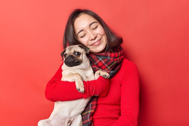 Gelukkig jonge Aziatische vrouw omarmt pug hond met liefde drukt liefde uit voor favoriete huisdier hebben vriendelijke relaties geniet van gezelschap van beste vriend draagt trui met sjaal geïsoleerd op rode achtergrond