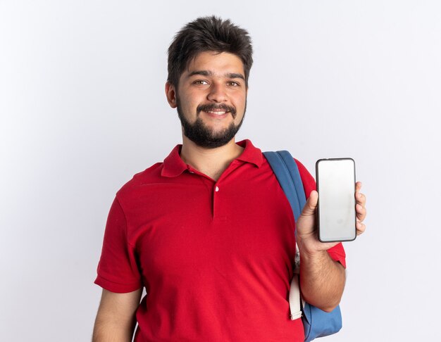 Gelukkig jonge bebaarde student man in rood poloshirt met rugzak smartphone glimlachend vrolijk staande op witte achtergrond tonen