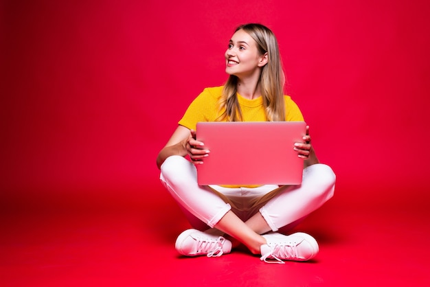 Gelukkig jonge krullend mooie vrouw zittend op de vloer met gekruiste benen en met behulp van laptop op rode muur.