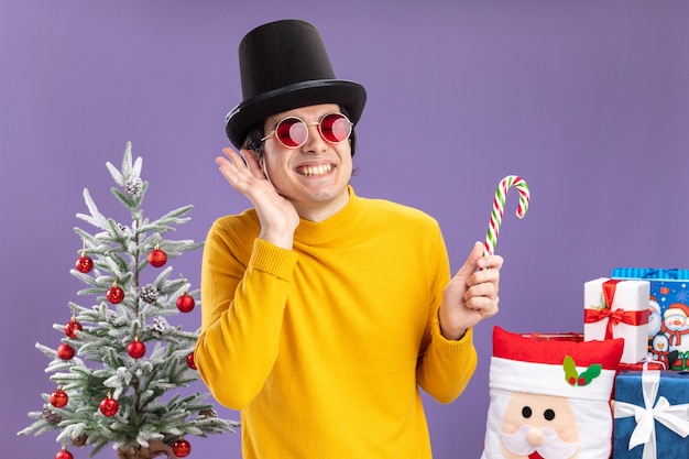 Gelukkig jonge man in gele coltrui en bril met zwarte hoed bedrijf candy cane glimlachend vrolijk permanent naast een kerstboom en presenteert op paarse achtergrond