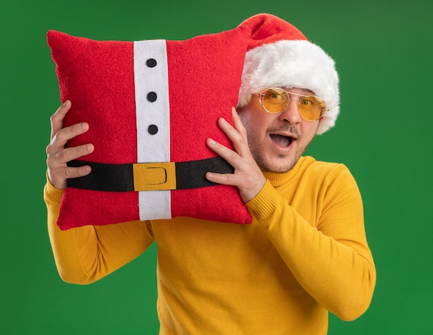Gelukkig jonge man in gele coltrui en kerstmuts met bril met rood grappig kussen kijken camera verrast staande over groene achtergrond