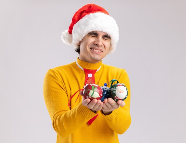 Gelukkig jonge man in gele coltrui en kerstmuts met kerstballen glimlachend vrolijk staande op witte achtergrond