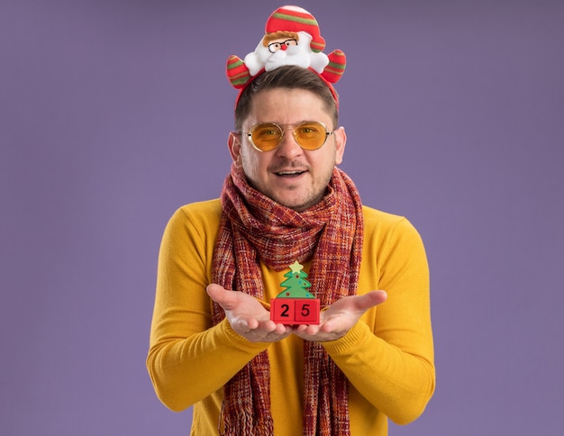 Gratis foto gelukkig jonge man in gele coltrui met warme sjaal en bril grappige rand met santa op hoofd weergegeven: speelgoed blokjes met nummer vijfentwintig staande op paarse achtergrond