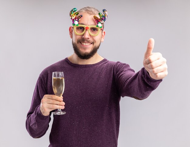 Gelukkig jonge man in paarse trui en grappige glazen houden glas champagne kijken camera met glimlach op gezicht schoen duimen omhoog staande op witte achtergrond
