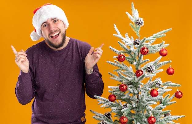 Gelukkig jonge man in paarse trui en kerstmuts camera kijken met glimlach op gezicht met opgeheven armen staande naast kerstboom over oranje achtergrond