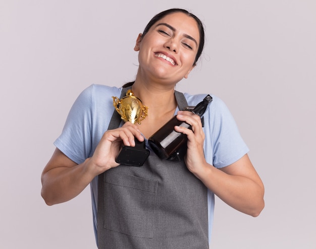 Gelukkig jonge mooie vrouw Kapper in schort met gouden trofee trimmer machine met spuitfles glimlachend met gesloten ogen staande over witte muur