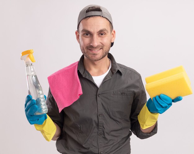 Gelukkig jonge schoonmaak man met vrijetijdskleding en pet in rubberen handschoenen met schoonmaakspray en spons met doek op zijn schouder glimlachend vrolijk staande over oranje muur