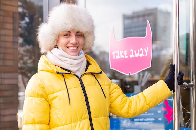 Gelukkig jonge vrouw aan de deur van het restaurant op een koude winterdag, belettering, afhaalmaaltijden.