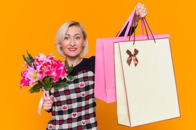 Gelukkig jonge vrouw in mooie jurk bedrijf boeket bloemen en papieren zakken met geschenken glimlachend vrolijk vieren internationale Vrouwendag staande over oranje muur