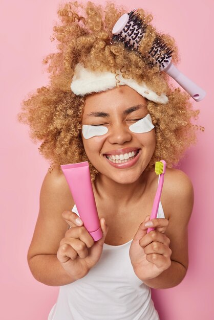 Gelukkig jonge vrouw met krullend borstelig haar houdt ogen gesloten toont perfecte witte tanden houdt tube tandpasta en tandenborstel draagt slaapmasker en casual wit t-shirt geïsoleerd over roze achtergrond