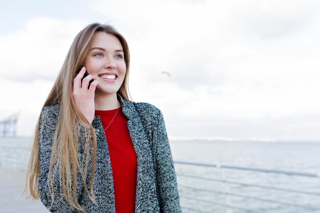 Gelukkig jonge vrouw met lang lichtbruin haar en prachtige glimlach praten over smartphone met prachtige glimlach in de buurt van de zee