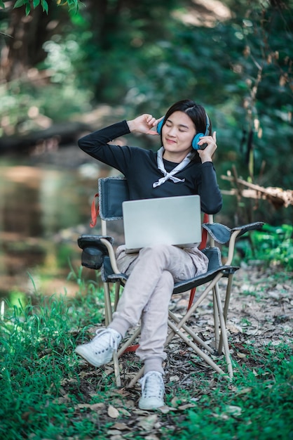 Gelukkig jonge vrouw zittend op een campingstoel met een koptelefoon die muziek luistert van een laptopcomputer terwijl ze ontspant op de camping in het bos