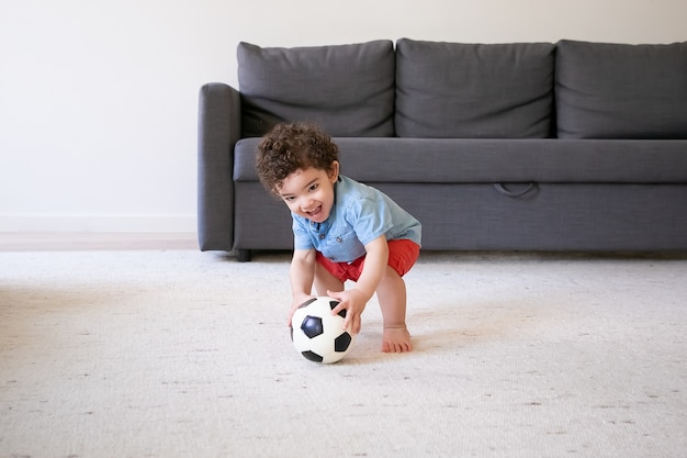 Gelukkig jongetje spelen met voetbal thuis, glimlachend. Schattige baby permanent op blote voeten tapijt en plezier in de woonkamer. Vakantie-, weekend- en jeugdconcept