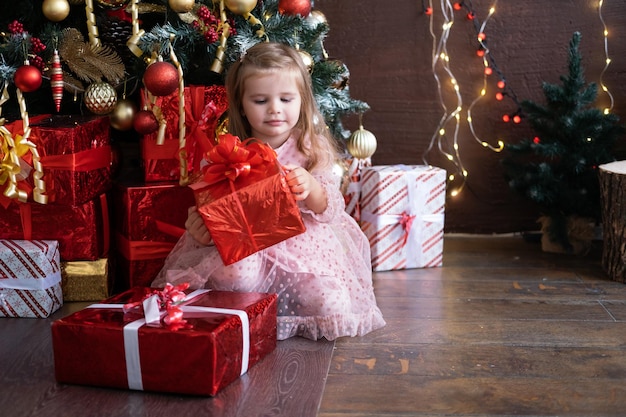 Gelukkig klein lachend blond meisje in mooie jurk met geschenkdoos in de buurt van kerstboom Premium Foto