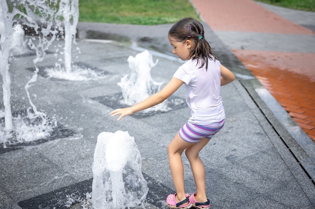 Gratis foto gelukkig klein meisje onder het opspattende water van de stadsfontein heeft plezier en ontsnapt aan de hitte.