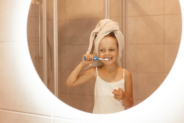 Gelukkig klein meisje tandenpoetsen in de badkamer, kijken naar haar spiegelbeeld in de spiegel, wit t-shirt dragen en haar haar in een handdoek wikkelen, ochtend hygiënische procedures.
